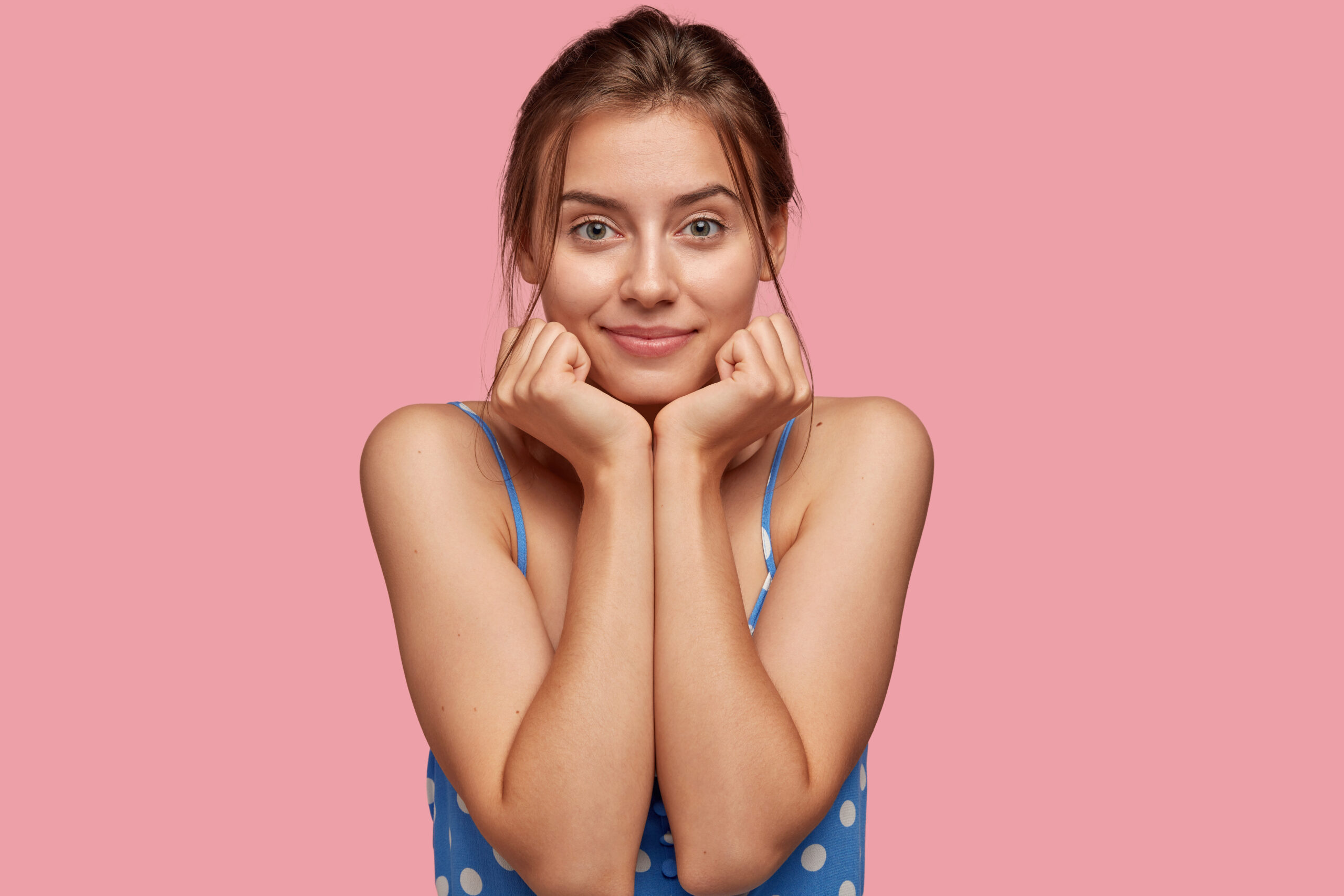 Charming beautiful female model keeps both hands under chin, looks seriously directly at camera, wears casual dress, has healthy skin, models against pink studio background. People and beauty concept